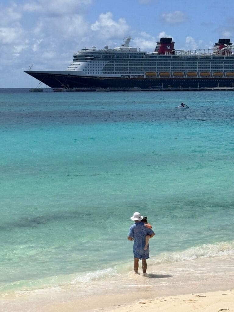 Person holding a child in shallow turquoise water with a Disney cruise ship docked in the background