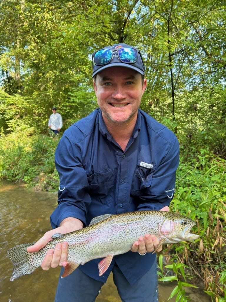 Man in a fishing shirt holding a rainbow trout beside a wooded stream