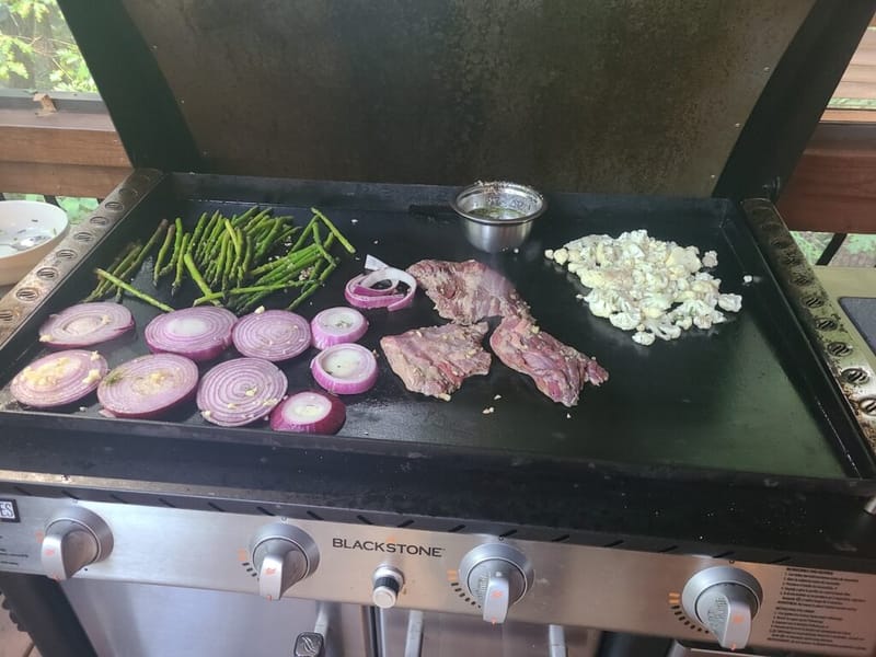 Steak, red onion, asparagus, and cauliflower cooking on an outdoor flat-top griddle